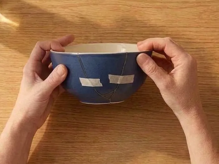 Two hands holding a navy bowl glued back together, strips of masking tape holding the seams while they cure